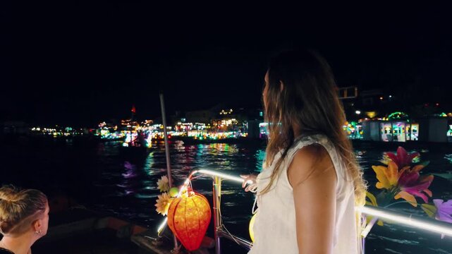 Young woman on a tourist boat ride admiring the colorful illuminated cityscape and traditional lanterns floating on the thu bon river at night, experiencing the local culture in hoi an, vietnam