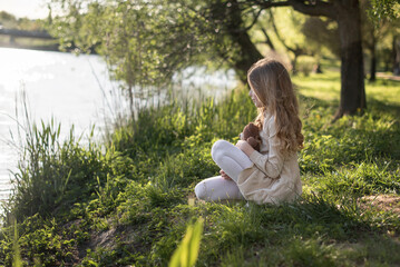 A young Caucasian girl with long brown hair sits by a riverbank, holding a teddy bear. She wears a light dress and enjoys the serene natural surroundings.