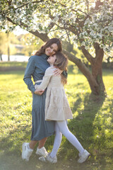 A young Caucasian woman embraces a young girl in a park. They stand under a flowering tree, enjoying a sunny day. Both wear casual spring outfits.