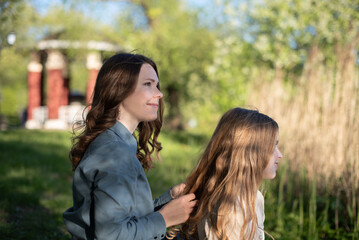 Two young Caucasian women in a park. One has long brown hair and is styling the hair of the other, who has long blonde hair. Green foliage surrounds them.