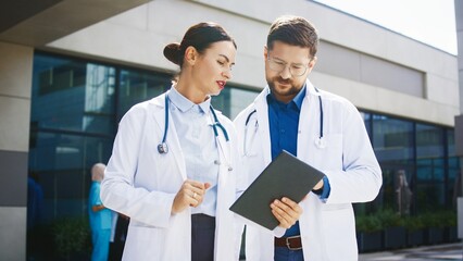 Caucasian female doctor standing outside clinic. Specialist discussing case details with male colleague holding tablet. Medical team focusing on shared evaluation. Outdoors near hospital building.