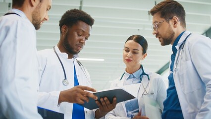 Focused male doctor holding chart while colleagues gathering around him in bright hospital hall. Reviewing patient notes. Clarifying duties. Coordinating tasks for busy clinical shift.