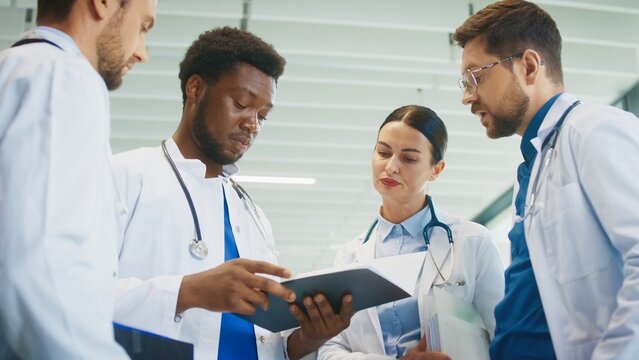 Focused male doctor holding chart while colleagues gathering around him in bright hospital hall. Reviewing patient notes. Clarifying duties. Coordinating tasks for busy clinical shift.