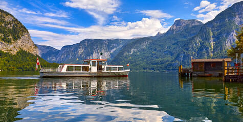Fototapeta premium Hallstatt, Austria. Touristic pleasure boat arriving to the old wooden dock at lake Hallstattersee among austrian Alps mountains. Picturesque summery landscape. Blue sky with clouds.