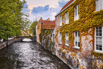 Fototapeta premium Bruges, Belgium. Medieval vintage brick houses with balconies over river with old stone bridge, covered with green plants and bushes. Summer landscape with sky and clouds..