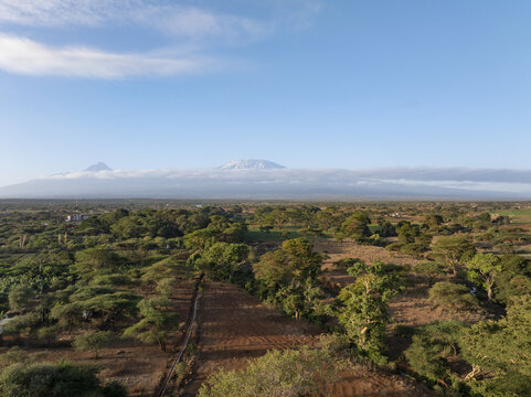 Aerial view of a vast, arid landscape dotted with verdant trees under a boundless sky, shadowed by distant, snow-capped peaks, Kimana, Kilimanjaro Region, Tanzania.