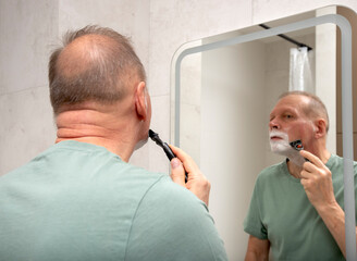A man shaves his neck and jawline while checking his reflection in the bathroom mirror at home.