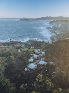 Aerial view of a cluster of futuristic, spherical white domes nestled amidst lush green vegetation, contrasting with the rugged coastline and the vast expanse of the ocean, Baredo, Pontevedra, Spain.