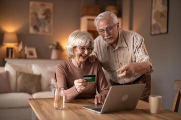 Cheerful Elderly Couple Shopping Online Together At Home With Laptop And Card