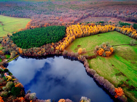 Aerial view of the lake's dark waters reflecting the sky, surrounded by vibrant autumn foliage and evergreen trees, creating a stunning contrast of colors, Morvan, France.