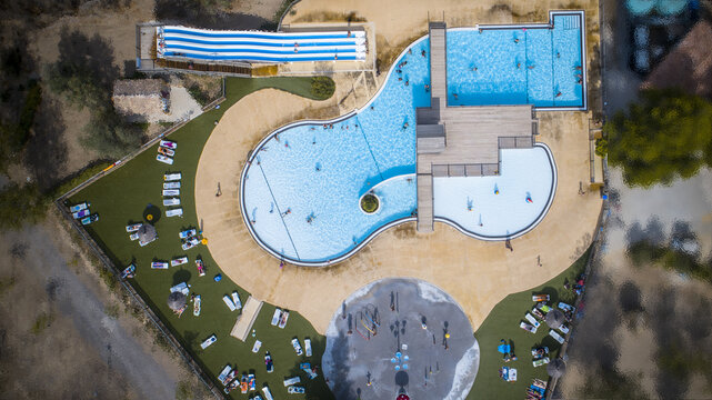 Aerial view of shimmering pools reflecting the azure sky, framed by sun-drenched lawns dotted with sunbathers, a vibrant oasis of leisure, Narbonne, France.