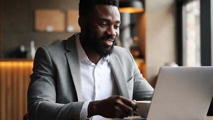 A businessman in a suit smiles while looking at a laptop and drinking coffee