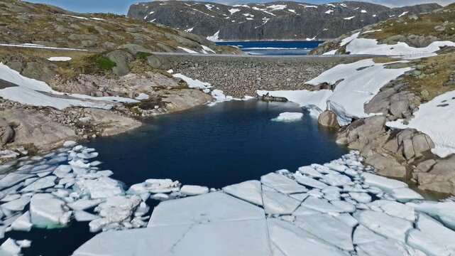 Mountain reservoir with melting ice near Hardangervidda Norway. Aerial view shows rocky shoreline, snow patches, and dark water breaking through ice.