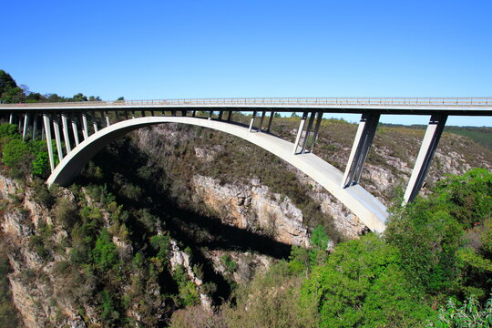 Concrete arch bridge spanning rocky gorge with green vegetation under clear blue sky