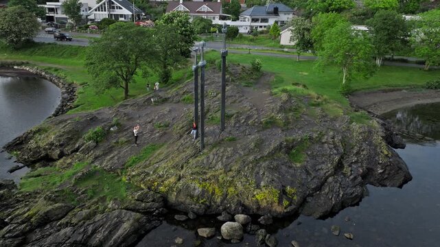 Low altitude drone flight above rocky island with Sverd i Fjell monument. Tourists stroll along the shore, discovering the Viking landmark during a calm visit.