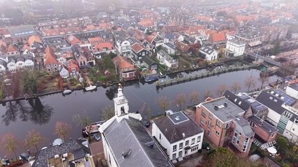 Aerial view of foggy Dutch canal town gabled brick homes, docked boats, and tall church tower form a tranquil, symmetrical waterfront scene softened by mist.