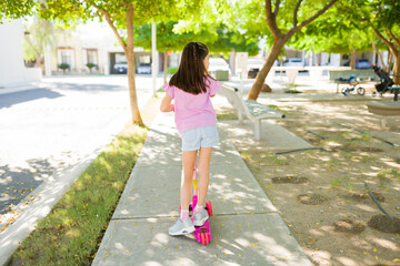 Rear view of girl riding scooter on park sidewalk during summer, enjoying outdoor playtime and a carefree childhood