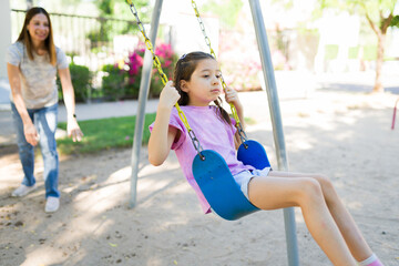Mother pushing daughter on a swing at a playground, child looking sad and experiencing childhood emotions