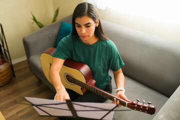 Latin woman learning to play acoustic guitar at home, practicing music with sheet notes in living room