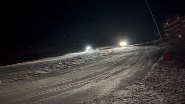 Two brightly illuminated snowcats move in opposite directions along ski slope