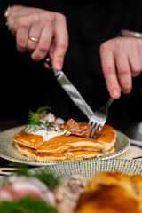 A close-up shot of hands using a knife and fork to cut into a stack of golden brown pancakes topped with sour cream, bacon bits, and fresh dill, served on a patterned plate.