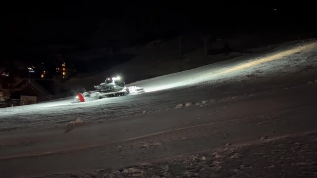 A brightly lit snowcat moves up the ski slope, compacting the snow beneath it