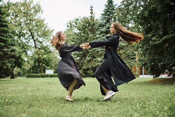 Two young women in long black coats dancing outdoors on green grass