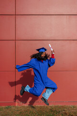African american student jumping with joy, holding a diploma and wearing a cap and gown