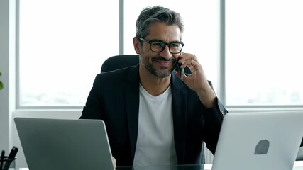 Cheerful Middle-Aged Businessman with Glasses and Beard Talking on Phone, Working with Laptops in Modern Office