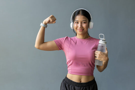 Confident Asian woman wearing headphones and holding water bottle while flexing arm. Concept of fitness success, healthy lifestyle, workout motivation, strength training, wellness mindset,