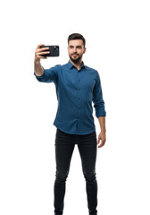 young handsome caucasian man with a beard, taking a selfie with a black smartphone, full body against a high-key bright white studio background. concept of modern social media lifestyle