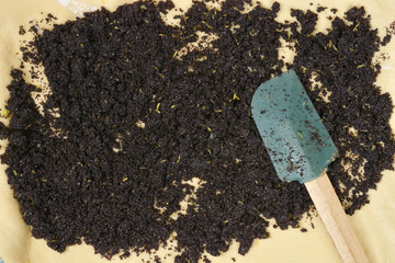 A top down close up shot of a dark poppy seed and sugar mixture being spread onto a thin layer of yeast dough with a green spatula