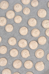 A high angle shot of several rows of unbaked beige french macaroon batter portions piped onto white parchment paper on a baking sheet