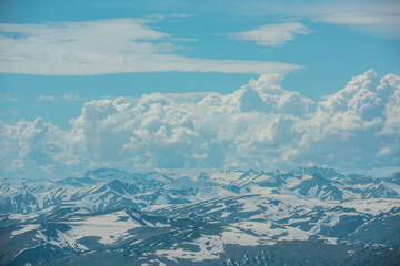 Aerial sunlit top view above snowbound hills and big snowy mountain range under lush clouds in changeable weather. Scenic spotty hilly alpine landscape. Partly cloudy in high snow-capped mountains.