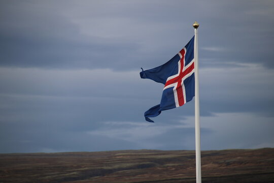 Icelandic flag on white pole against cloudy sky and hilly landscape