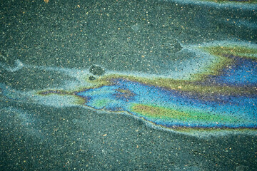 A multicolored puddle of gasoline shimmers with bright iridescent reflections on textured dark asphalt.