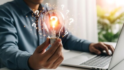 Person holding illuminated incandescent light bulb featuring a glowing question mark surrounded by floating query symbols while working on a laptop.