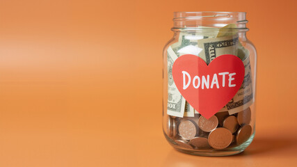 Charity Donation Jar &ndash; Glass Container with Coins, Heart Label, and Orange Background for Fundraising Concepts and Financial Aid Campaigns