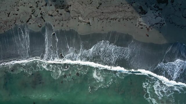 Top-down drone aerial of powerful ocean waves breaking on black volcanic sand beach with white foam and dramatic surf patterns