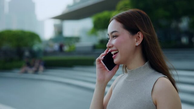 Portrait of happy Asian woman talks on smartphone as walks across urban square slow motion. Young brunette Korean female calls girl friend by mobile phone in megapolis district