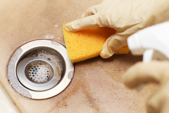 Cleaning kitchen sink drain with yellow sponge. Gloved hands scrubbing metal strainer area. Removing dirt and stains around drain hole. Home hygiene routine. Household cleaning process.