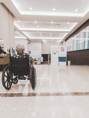 Rear view of a senior woman sitting in a wheelchair inside a modern hospital lobby with bright lighting and spacious interior. Healthcare concept representing elderly care, patient support, accessibil