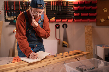 A professional carpenter works with wood using carpentry tools in her garage, taking orders over the phone and writing them down in a notebook. Craft, creative small business