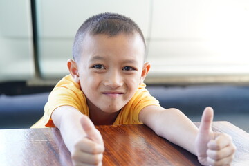 Happy young boy giving double thumbs up while sitting at a wooden table with a confident smile, expressing joy and positivity in daily life moments