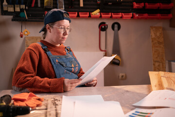 professional female carpenter works with wood using carpentry tools in her garage. She sits, holds sheets of paper with graphs, and studies the material. Concept of profession, art, and hobby.