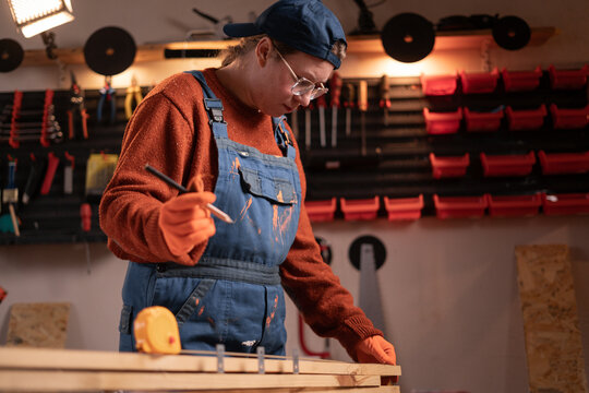 A female carpenter works in her garage, using professional woodworking tools. Handicrafts, creativity, and carpentry. She makes marks on a piece of wood with a simple pencil - Powered by Adobe