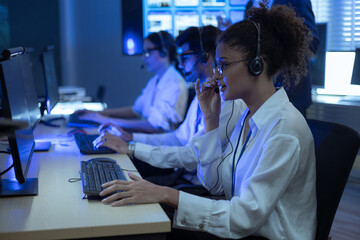 Diverse customer support team with headsets working in a dark modern office. Professional technical agents providing service and telemarketing solutions using computers at night shift