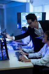 Professional diverse IT support team working in a dark technical control room. A male manager uses a stylus to point at a monitor while guiding a female operator wearing a headset at her desk