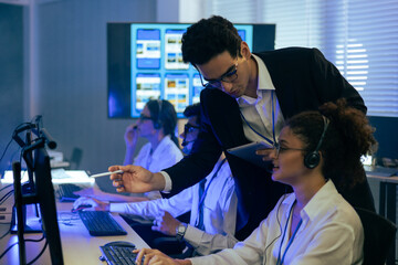 Professional manager supervising a diverse customer support team with headsets in a modern office. He points at a computer screen using a digital tablet to guide staff in a technical center.