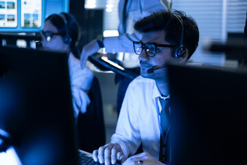 Focused male security operator with headset monitors data on screens in a high-tech control room. Professional team managing cybersecurity, network safety, and digital system operations daily